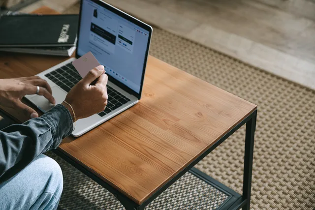 Person holding a credit card while shopping online using a laptop on a wooden table. Ecommerce conversion channels: one shared record for web or seller app, marketplace and assisted routes