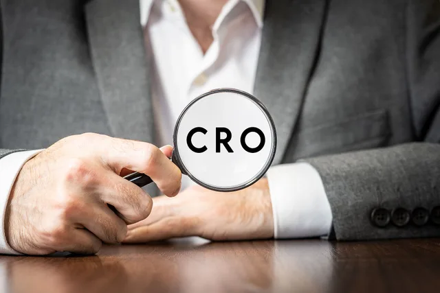 An image of a man holding up a magnifying glass with the acronym ‘CRO’ inside the glass. The man is wearing a grey suit and a white shirt, and is leaning on a desk in the background. ‘CRO’ is short for the ecommerce term ‘conversion rate optimisation’. This image is free to use for any purpose under. Selecting conversion suppliers by one store job, one dossier and independent risk gates